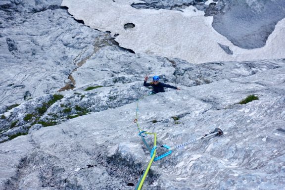 Kletterer besteigt steile Felsen mit Seil und Sicherungsgerät, Umgebung aus grauem Gestein.