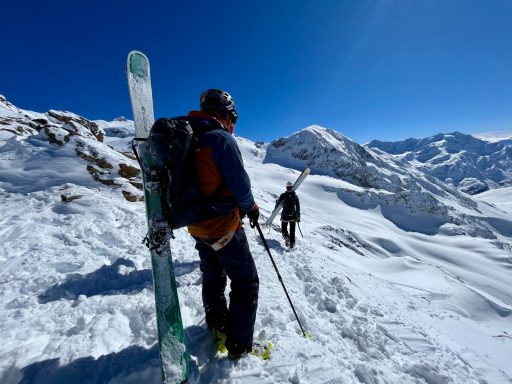 Zwei Skifahrer in verschneiter Berglandschaft, einer mit Skiern auf dem Rücken.