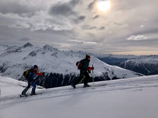 Zwei Personen wandern im Schneegebirge unter einem bewölkten Himmel.