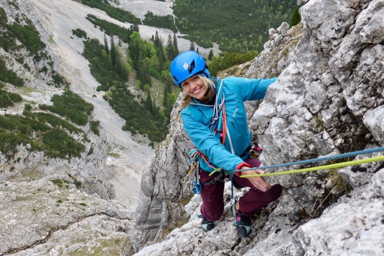 Eine bergsteigende Person in blauer Ausrüstung lächelt, umgeben von felsiger Landschaft.