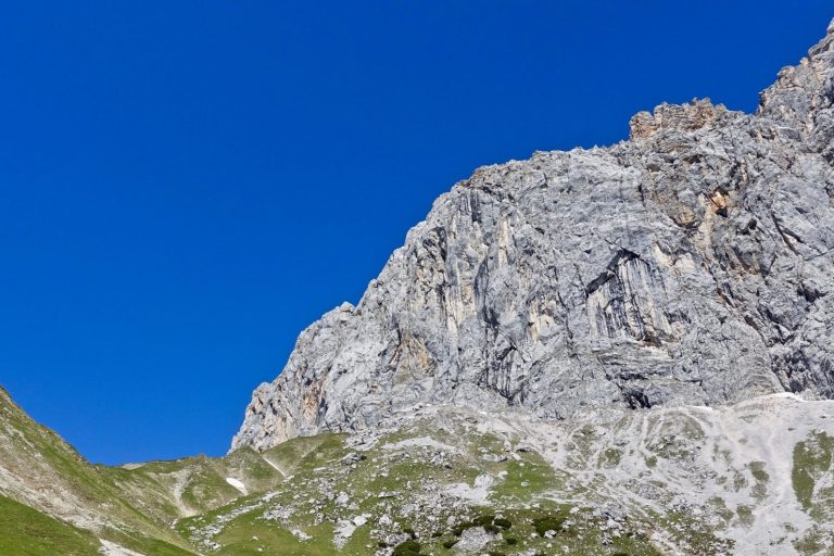 Hochalpine Felsen unter klarem, blauem Himmel mit grünen Wiesen im Vordergrund.