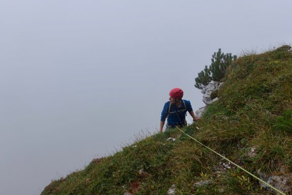 Ein Kletterer mit rotem Helm erklimmt eine steile, grasbewachsene Felswand.