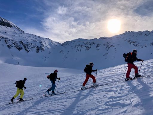 Vier Skifahrer steigen einen verschneiten Hang unter einem sonnigen Himmel hinauf.