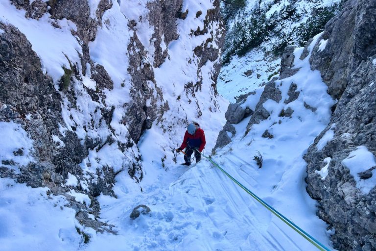 Bergsteiger in roter Jacke klettert durch eine schneebedeckte Schlucht.