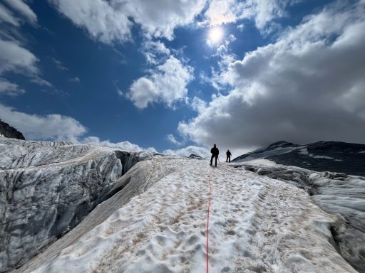 Zwei Personen wandern über einen schneebedeckten Gebirgsweg unter einem bewölkten Himmel.