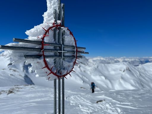 Schneebedeckter Berggipfel mit einem markierten Wegweiser und einer Person im Hintergrund.