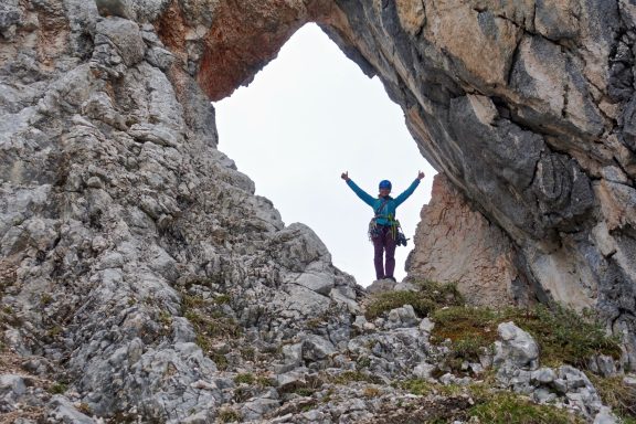 Bergsteiger steht triumphierend in einem Felsenbogen mit erhobenen Armen.