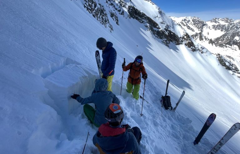 Drei Personen im Schnee, eine gräbt, zwei stehen und genießen die winterliche Berglandschaft.