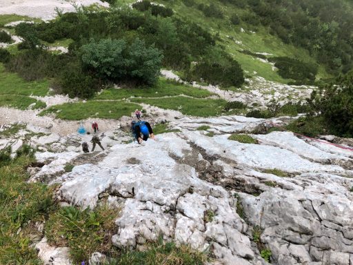 Zwei Personen klettern auf einem steinigen Hang in einer grünen, bergigen Landschaft.