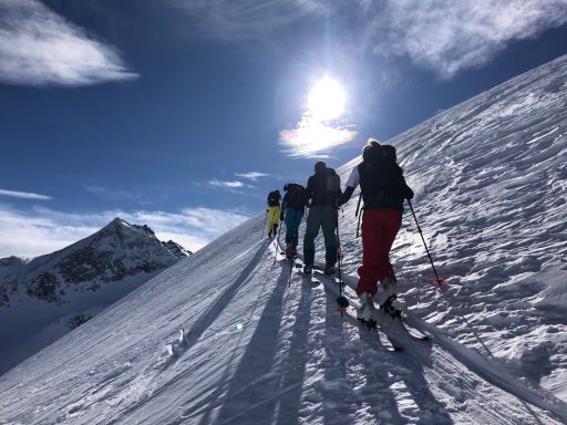 Skifahrer steigen einen schneebedeckten Hang hinauf, sonniger Himmel im Hintergrund.
