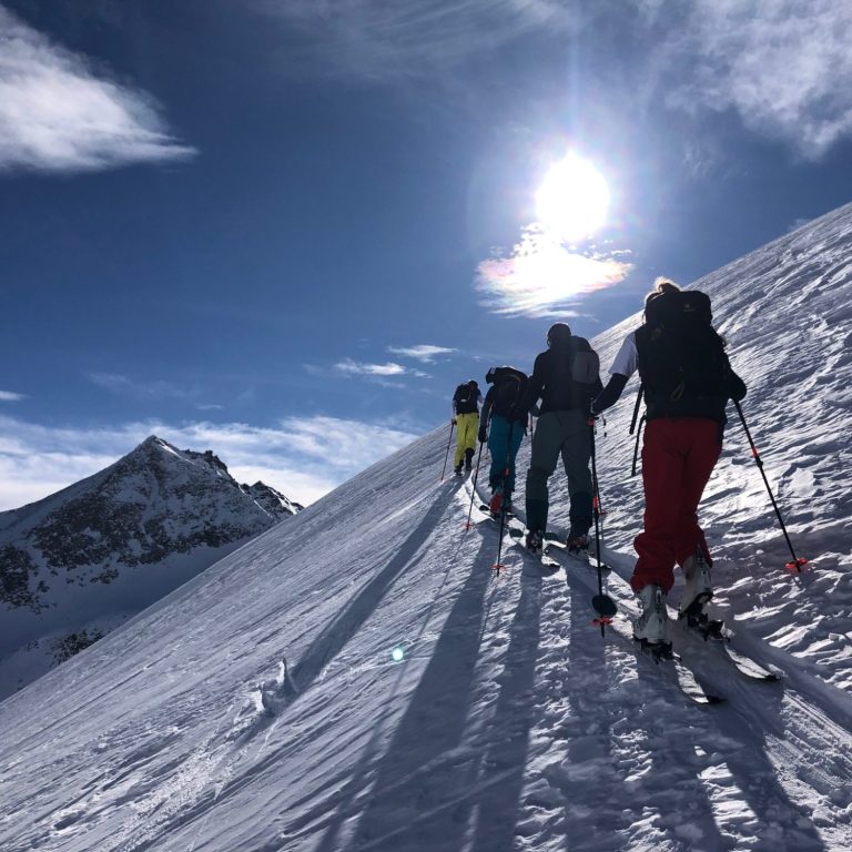 Skifahrer steigen einen schneebedeckten Hang mit Sonnenschein und Bergen im Hintergrund hinauf.