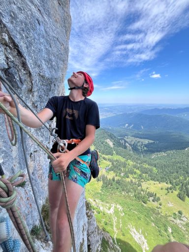Kletterer in Berglandschaft, schaut nach oben, gesichert mit Seil und Kletterausrüstung.