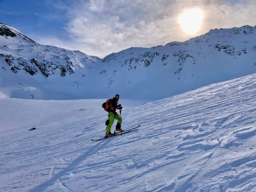 Skifahrer auf verschneiter Piste mit Bergen und Sonne im Hintergrund.