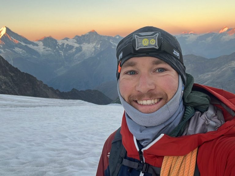 Mann in warmer Kleidung, lächelt vor schneebedeckten Bergen bei Sonnenaufgang.