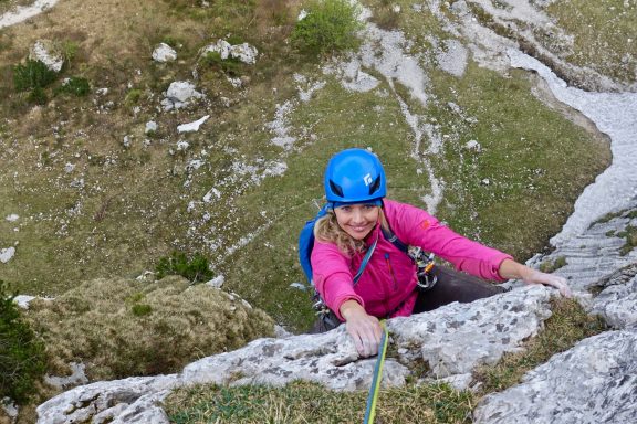 Dame climber in pink shirt and blue helmet klettert an einer Felswand hoch.