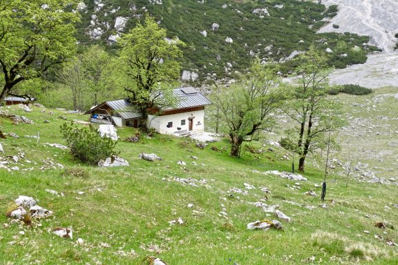 Ein einfaches Haus umgeben von grünen Wiesen und Bäumen in einer bergigen Landschaft.