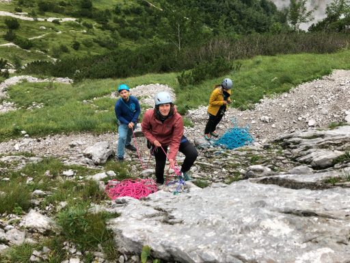 Drei Personen klettern in der Natur, mit bunten Seilen und Helmen, umgeben von Felsen.