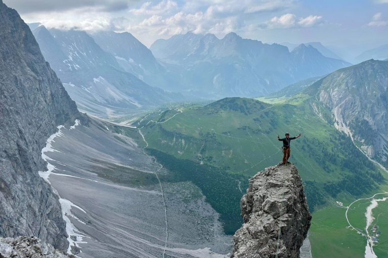 Junge Frau klettert an einer Felswand, gesichert mit einem Seil, auf einem grünen Berg.