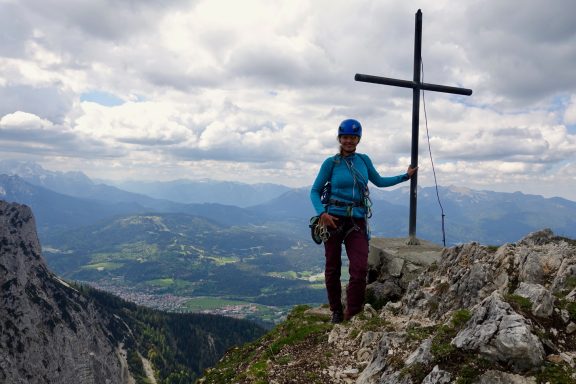 Person steht neben einem Kreuz auf einem Berg, mit einer weiten Landschaft im Hintergrund.