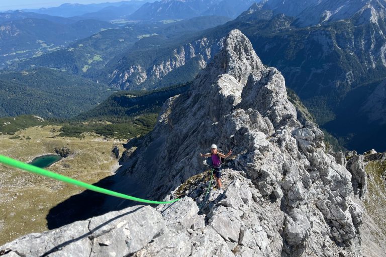 Person steht auf einem schmalen Felsgrat mit Seil und Blick auf die Berge.