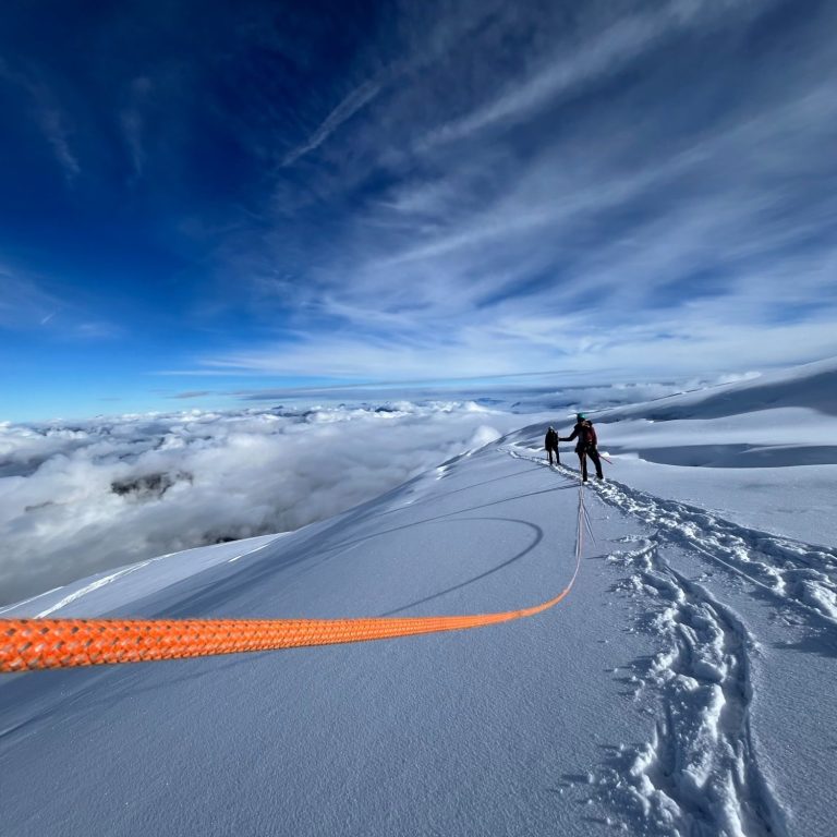 Zwei Personen wandern auf einem verschneiten Gipfel unter blauem Himmel.