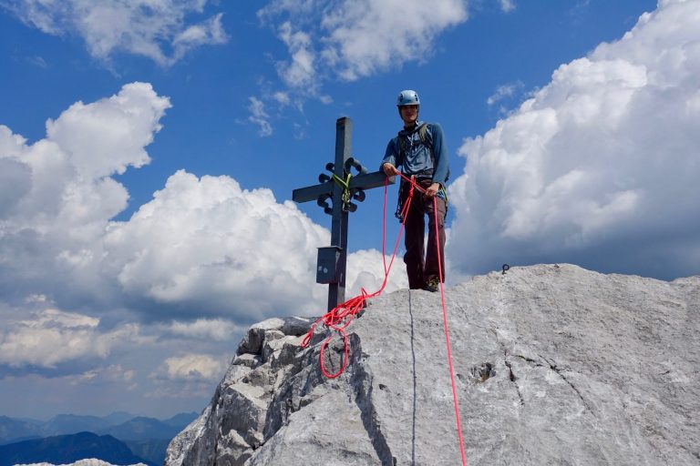 Bergsteiger mit Seil steht auf einem Gipfel unter einem Kreuz, umgeben von Wolken.