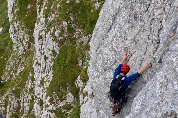 Kletterer in roter Helm an steiler Felswand mit grüner Landschaft im Hintergrund.