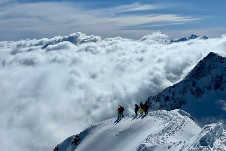 Menschen stehen auf einem Gipfel, umgeben von Wolken und schneebedeckten Bergen.
