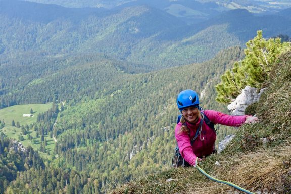 Frau klettert an einer Felswand mit Blick auf eine grüne, bewaldete Landschaft.