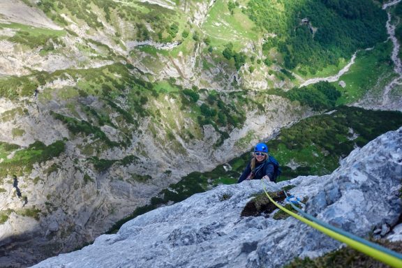 Kletterer an einer Felswand, mit Blick auf ein tiefes Tal und grüne Berghänge.