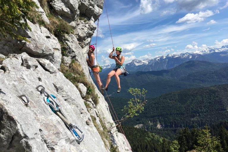 Zwei Kletterer hängen an einer Felswand mit Berglandschaft im Hintergrund.