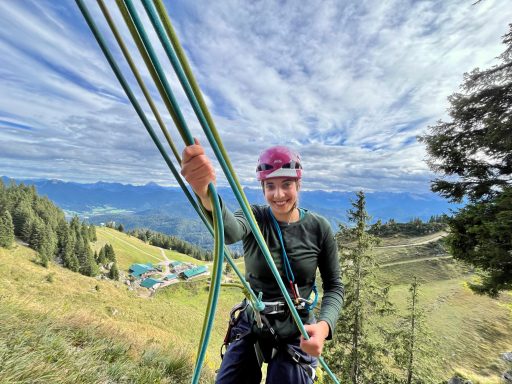 Person beim Klettern, hält Seile mit Bergblick und wolkenverhangenem Himmel im Hintergrund.