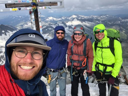Vier Bergsteiger posieren lächelnd am Gipfel, mit Aussicht auf schneebedeckte Berge.