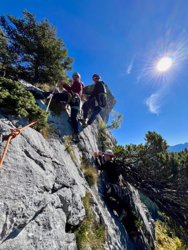Drei Kletterer an einer Felswand unter blauem Himmel und Sonnenschein.