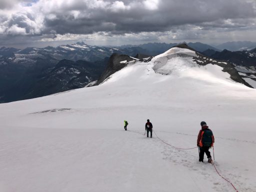 Drei Bergsteiger am Hang eines schneebedeckten Berges unter bewölktem Himmel.