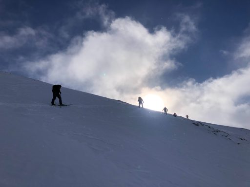 Schneebedeckte Hügel mit Menschen, die einen Hang hinaufsteigen, im Hintergrund Sonne und Wolken.