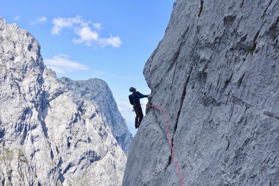 Bergsteiger klettert an einer steilen Felswand mit blauem Himmel im Hintergrund.