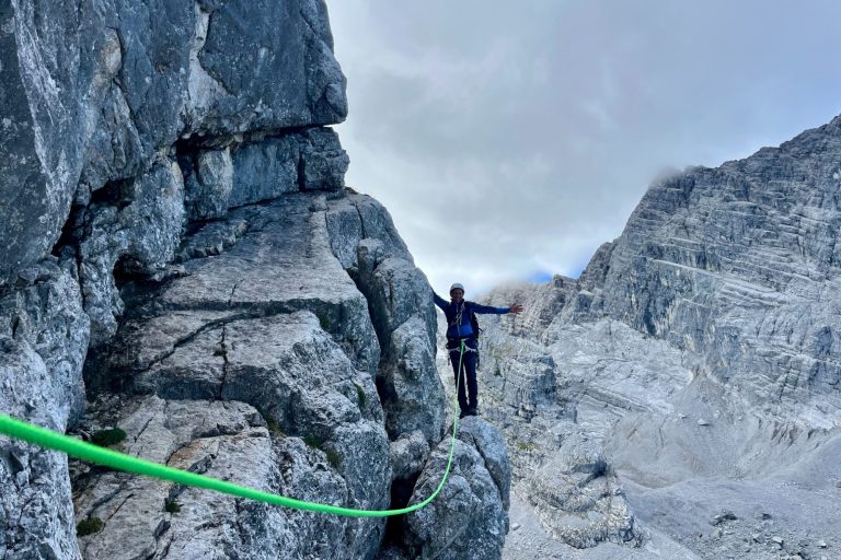 Kletterer balanciert auf einem schmalen Felsenhang mit Seilsicherung in bergiger Landschaft.