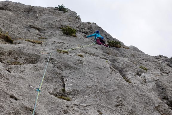 Kletterer in blauer Jacke an einer felsigen Wand, gesichert durch ein Seil.