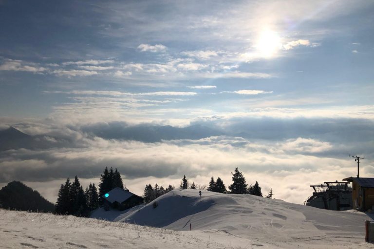 Berglandschaft mit Schnee, Bäumen und Wolken unter einem sonnigen Himmel.