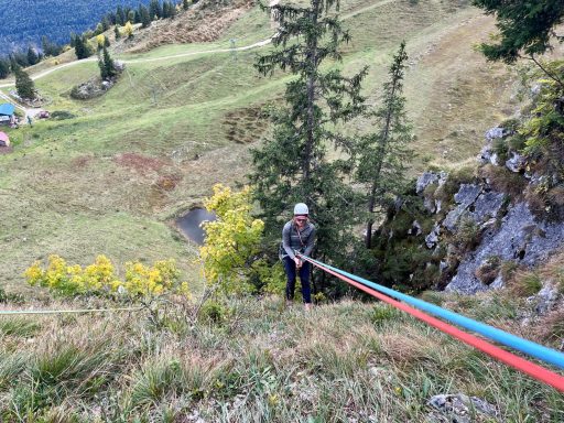 Bergsteiger sichert sich beim Abseilen an einer steilen Wiese mit Seil und Helm.