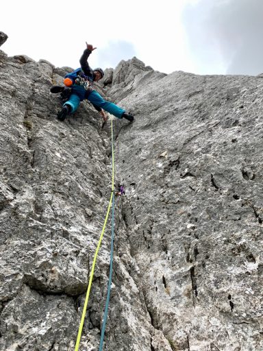 Kletterer an steiler Felswand mit Seil und Ausrüstung, Hände in der Luft.
