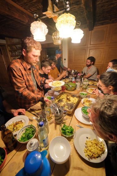 Skitouren Camp Rofan Ein Mann serviert Essen an einem gedeckten Tisch mit mehreren Gästen.